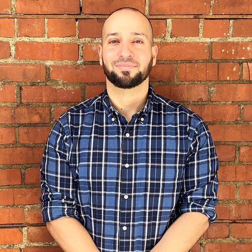 A man in a blue plaid shirt with facial hair standing against a brick wall, giving his exit interview from the youth program, MyComcle.